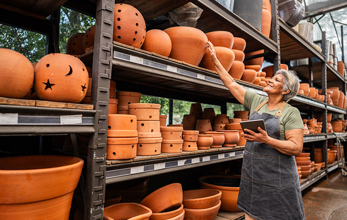 Woman wearing apron standing in an ourdoor store setting is observing shelves stocked with terracotta planting pots and garden decorations.