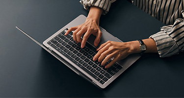 Close up image of two hands poised and typing over a laptop keyboard over a dark grey solid surface